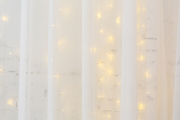 New Year or Christmas background. Greeting card. White tulle curtain on the background of a white brick wall decorated with a burning garland.