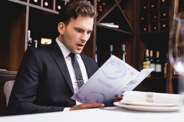 Selective focus of shocked man in formal wear looking at menu in restaurant