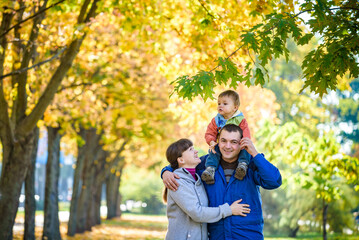Fototapeta premium Family of three walks in the autumn park holding hands. happy father carrying son with maple leaves. Mother embrace her cute boy child. Happy family leisure together concept