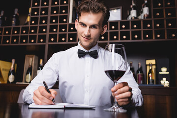 Selective focus of sommelier looking at camera while holding glass of wine and writing on notebook in restaurant