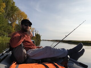 trolling fishing, a bearded man on a motor boat floating on the river on an autumn Sunny day and drinking tea from an iron mug.