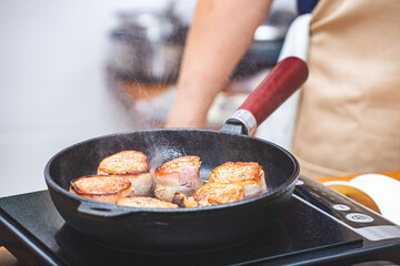 Meat wrapped in bacon is fried in a cast-iron skillet on the hotplate, a male chef wearing an apron in the background.