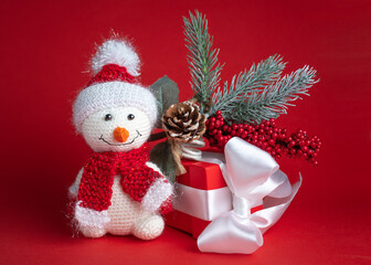 A snowman toy in a Santa Claus hat on a red background next to a red gift box tied with a white ribbon and an evergreen spruce twig with frost, cones and berries
