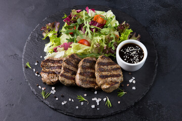 Grilled beef meat with sauce and salad of herbs, arugula, cherry tomatoes on a black board on a black background. Background image, copy space