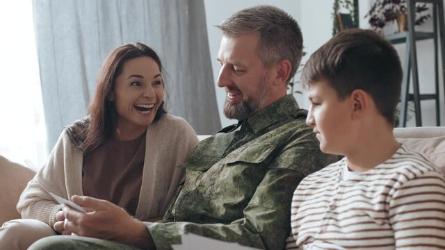 Handheld slowmo shot of cheerful male army veteran with beard sitting on couch and sharing deployment stories while showing photos to his happy wife and son