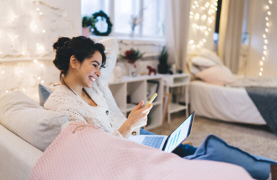 Laughing Woman Using Smartphone And Laptop On Sofa In Cozy Apartment