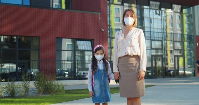 Joyful beautiful Caucasian woman standing in mask with little daughter outdoors. Happy mother holding hand of cute schoolgirl at schoolyard on street in good mood. Back to school. Quarantine concept