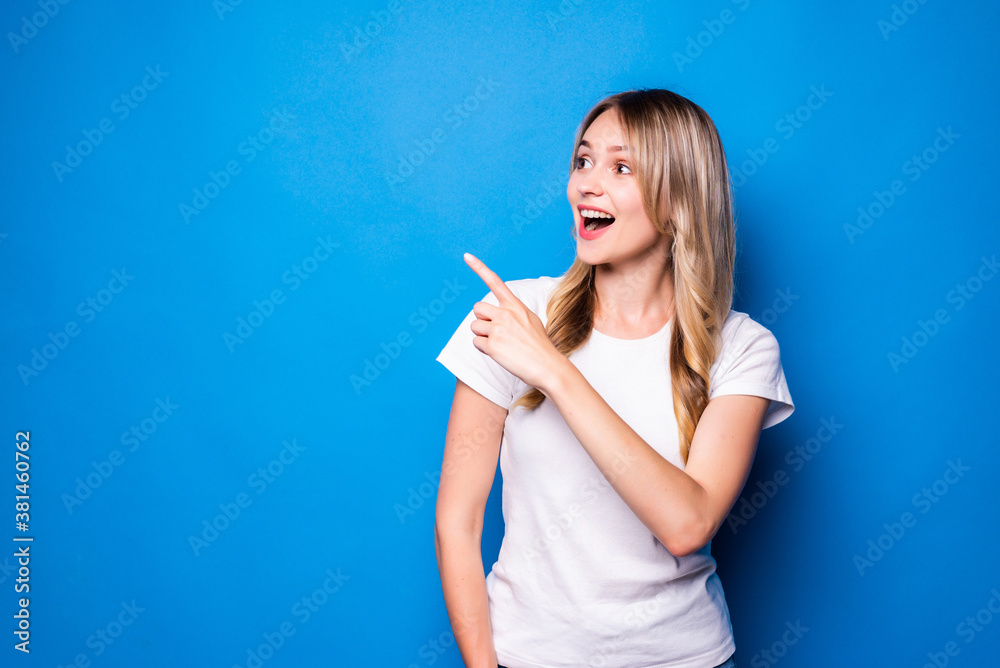 Wall mural Young woman pointing finger to the side over isolated blue background