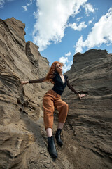 Fashion red-Haired girl in orange jeans posing in nature near sandy rocks, not like everyone else