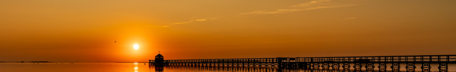 Fototapeta premium Large wooden pier on a beach just after sunset, bridge is in black silhouette and the sky of saturated with warm orange tones from the sunset.