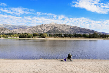 Obraz na płótnie Canvas Fisherman angling in the lakeshore of the “Santillana” reservoir with the “Guadarrama” mountain range in the background