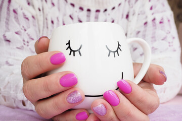 female hands with beautiful lilac manicure hold a white mug with a painted muzzle..