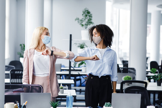 New normal and hello or hi, colleague. Millennial european and african american women in protective masks, greet with elbows in morning in interior of coworking office
