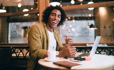 Portrait of prosperous happy curly male freelancer enjoying remote job making research online on laptop computer, young handsome 20s man student enjoying learning courses via technology in cafeteria