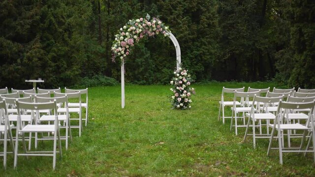 Beautiful wedding floral decorations of flowers in pastel faded colors, wooden painted white frame for outside wedding ceremony in park, raws of many empty chairs standing on green grassy lawn.