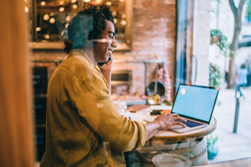 Successful businessman in casual wear typing on laptop computer making booking during smartphone call, positive young male freelancer enjoying remote job and mobile phone conversation in cafe