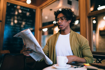 Serious concentrated male in eyewear spending time in cafe interior reading news from printed media on leisure, handsome curly man in casual wear holding newspaper checking financial information