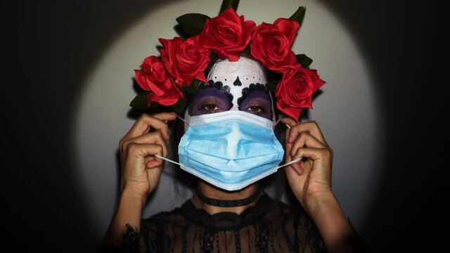 Concepto de Carnaval o Halloween. Mujer asustada con mascarilla anti covid y virus de la pandemia, pintada de calavera mexicana blanca, usa ropa negra en un escenario de miedo 4K
