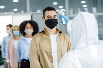 Fototapeta na wymiar Checking temperature in morning before work. Man in hazmat suit checks first symptoms of illness in workers in protective masks in office