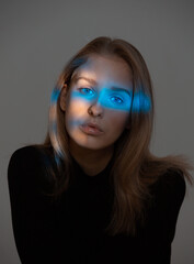 Fototapeta premium Portrait of young pretty girl with blue neon light on her face posing in studio on gray background