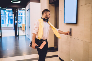 Smart casual coworker with modern digital tablet press button waiting for elevator for level up in office workspace, millennial businessman holding technology in corporate hall with blank copy space