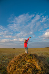 A girl with long hair in a field on a haystack enjoys the sun with her hands raised