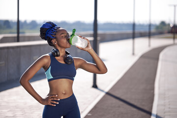 portrait of sporty african woman drinking water from bottle, sweaty female stand after running,...