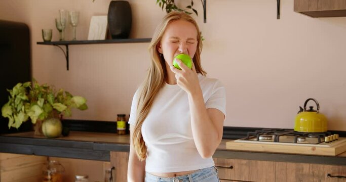 Young blonde woman is throwing up green apple and eating it in the kitchen. Healthy useful via harmful nutrition, diet concept, eating disorder. Happy fresh organic lifestyle with good food. 