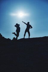 Dark silhouettes of a young couple of a guy and a girl of travelers on the top of the mountain indulge and rejoice against the background of the sun