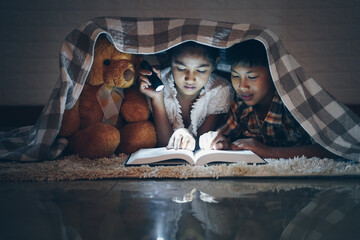 Happy children boy and girl reading a book in blanket at home with a toy teddy bear.