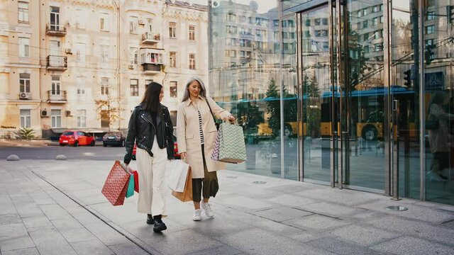 Asian mature female and adult daughter are smiling, talking, carrying colorful shopping packages, walking along glass facade building, slow motion