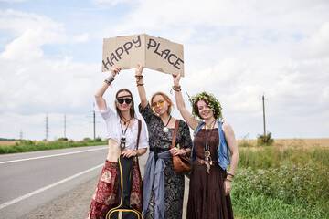 Three hippie women, wearing boho style clothes, standing on road, thumbing a ride, hitchhiking with sign Happy place on cardboard. Friends, traveling together in summer. Freedom and happiness concept.