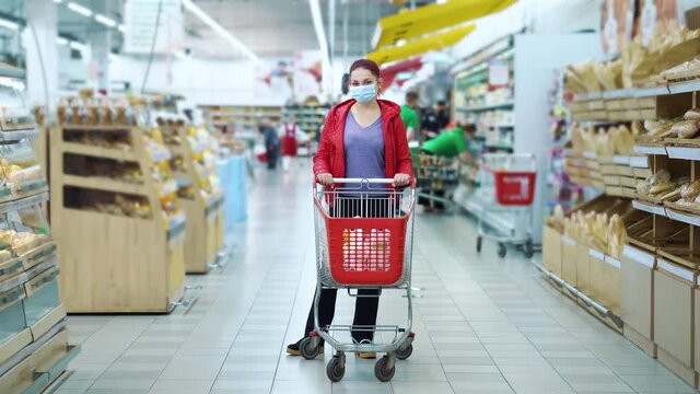 Young woman in medical mask standing with shopping cart between aisles in supermarket and looking at camera, blurred workers in uniform on background. Customer buying food during pandemic