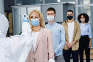 Temperature control test at work and social distancing. Man in protective suit checks multinational workers with non-contact thermometer in morning