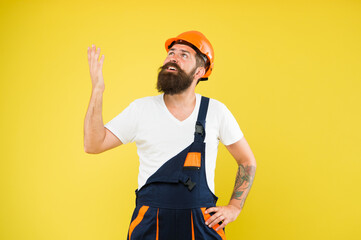 Bearded builder in hard hat and work uniform look up yellow background, construction