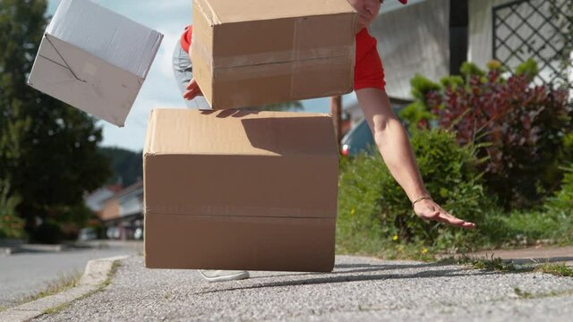 SLOW MOTION, LOW ANGLE, CLOSE UP, DOF: Clumsy Caucasian delivery man falls to the ground and drops cardboard boxes. Young male courier trips and falls to the asphalt pavement along with a few parcels.
