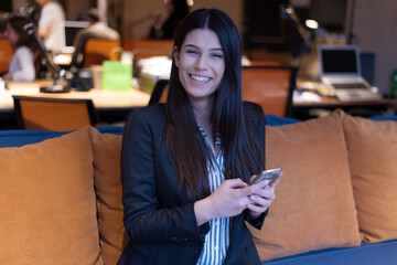 Young American business woman sitting at her sofa, using phone in her office and smiling.