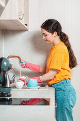 Housework. A pretty woman in casual clothes and pink rubber gloves is washing dishes. Side view