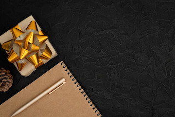 One gift wrapped in a craft paper and a golden ribbon. A notepad, a pen and a pine cone on black concrete background. Copy space, top view, flat lay.