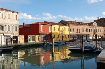 Fototapeta na wymiar Venetian's island of Burano, view of canal and traditional coloured houses facades, bright and dark sky after a storm, reflection on the canal with bridge and water ponds on the street, Venice view.