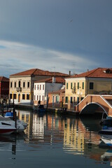 Obraz premium Venetian's island of Burano, view of canal and traditional coloured houses facades, bright and dark sky after a storm, reflection on the canal with bridge and water ponds on the street, Venice view.