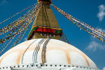 The upper part of the Boudhanath stupa in Kathmandu, Nepal. The image of Buddhas eyes on a gold surface of the stupa. Colorful prayer flags nearby.