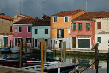 Venetian's island of Burano, view of canal and traditional coloured houses facades, bright and dark sky after a storm, reflection on the canal with bridge and water ponds on the street, Venice view.