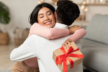Fototapeta na wymiar Joyful Asian Girlfriend Hugging Boyfriend Holding Wrapped Gift Box Indoors