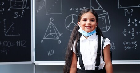 Portrait of cute happy small Caucasian girl with backpack standing in classroom, looking at camera and taking off medical mask. Little Hispanic schoolgirl smiling in school. Coronavirus.