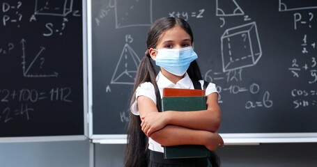 Portrait of cute small Caucasian pretty girl in medical mask and with backpack standing in classroom, looking at camera and holding textbooks. Little schoolgirl at blackboard in school. Pandemic time.