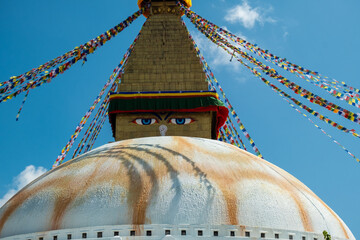 The upper part of the Boudhanath stupa in Kathmandu, Nepal. The image of Buddhas eyes on a gold surface of the stupa. Colorful prayer flags nearby.
