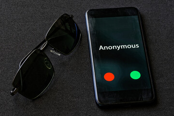 A black smartphone with an anonymous call lies on a dark fabric surface next to sunglasses. The...