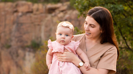 Happy young mother holds her young baby daughter in her arms outdoors. Mom smiles and looks at her daughter.