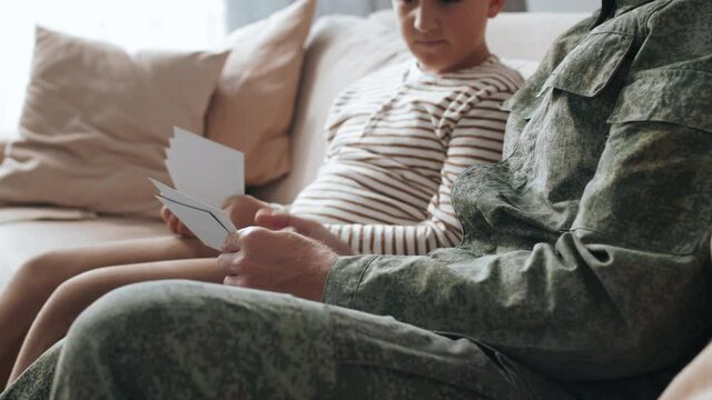 Tilt up shot of bearded male army veteran in uniform sitting on couch and showing photos to his son while sharing deployment stories